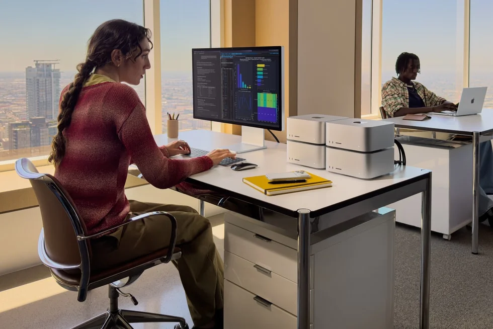 Two stacks of Mac Studio computers on an office desk beside a monitor showing charts, with the city visible through the window.